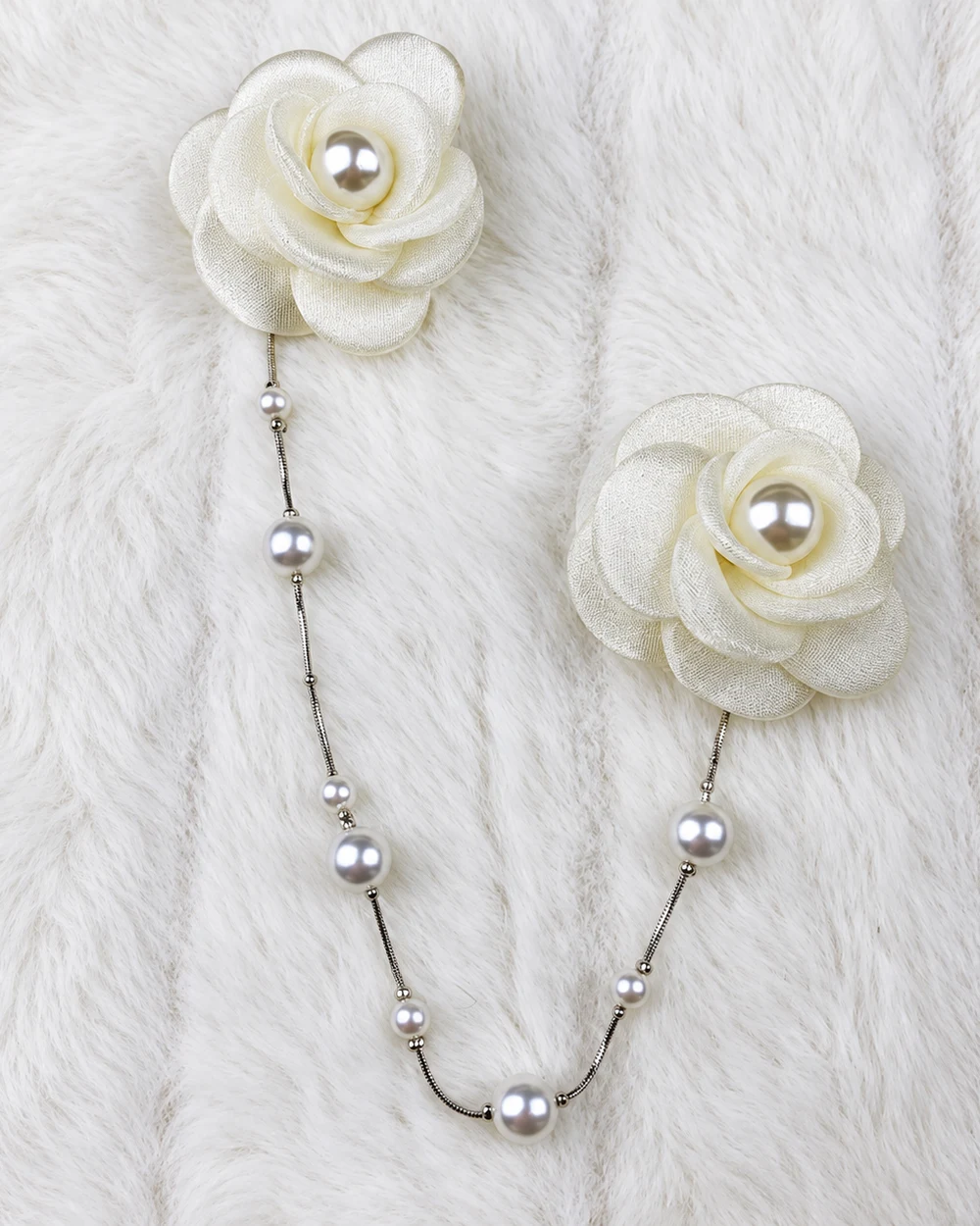 Two ivory fabric camellias with pearl-look centres connected by a silver bead chain on a soft white background.