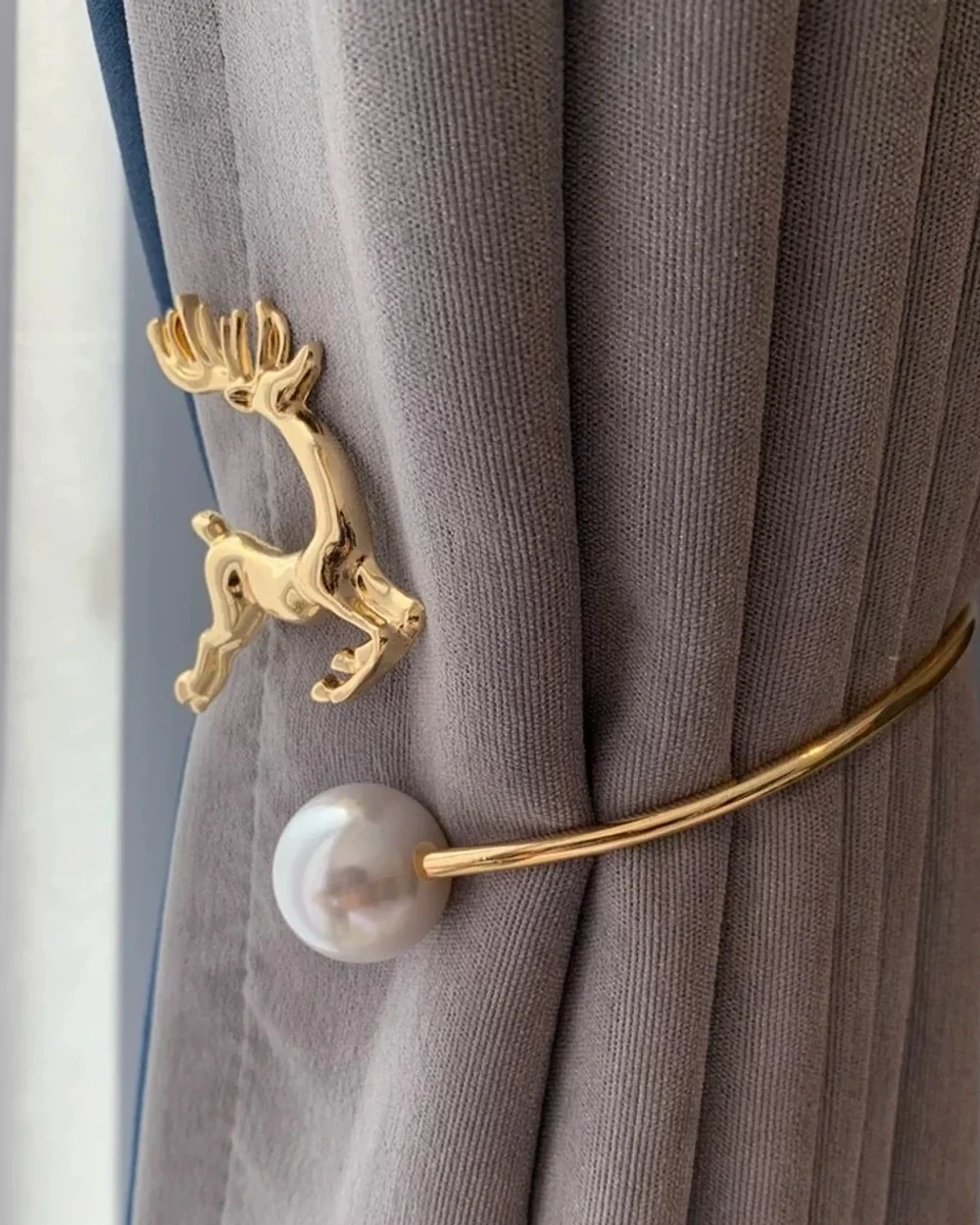 Gold-tone stag and a large pearl-look sphere at the ends of a curved tieback around a grey curtain.