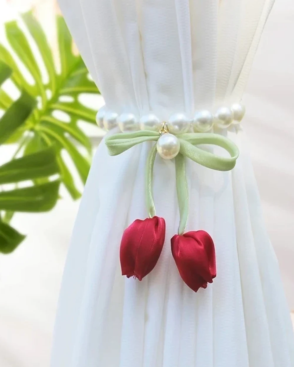 Two red fabric tulip buds hanging from a pale green ribbon bow and pearl-look beaded band around a white curtain.