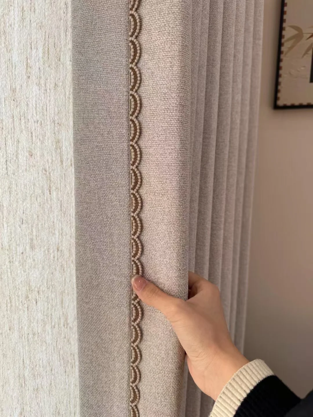 Hand holding beige-grey curtain edge with a scalloped decorative braid.