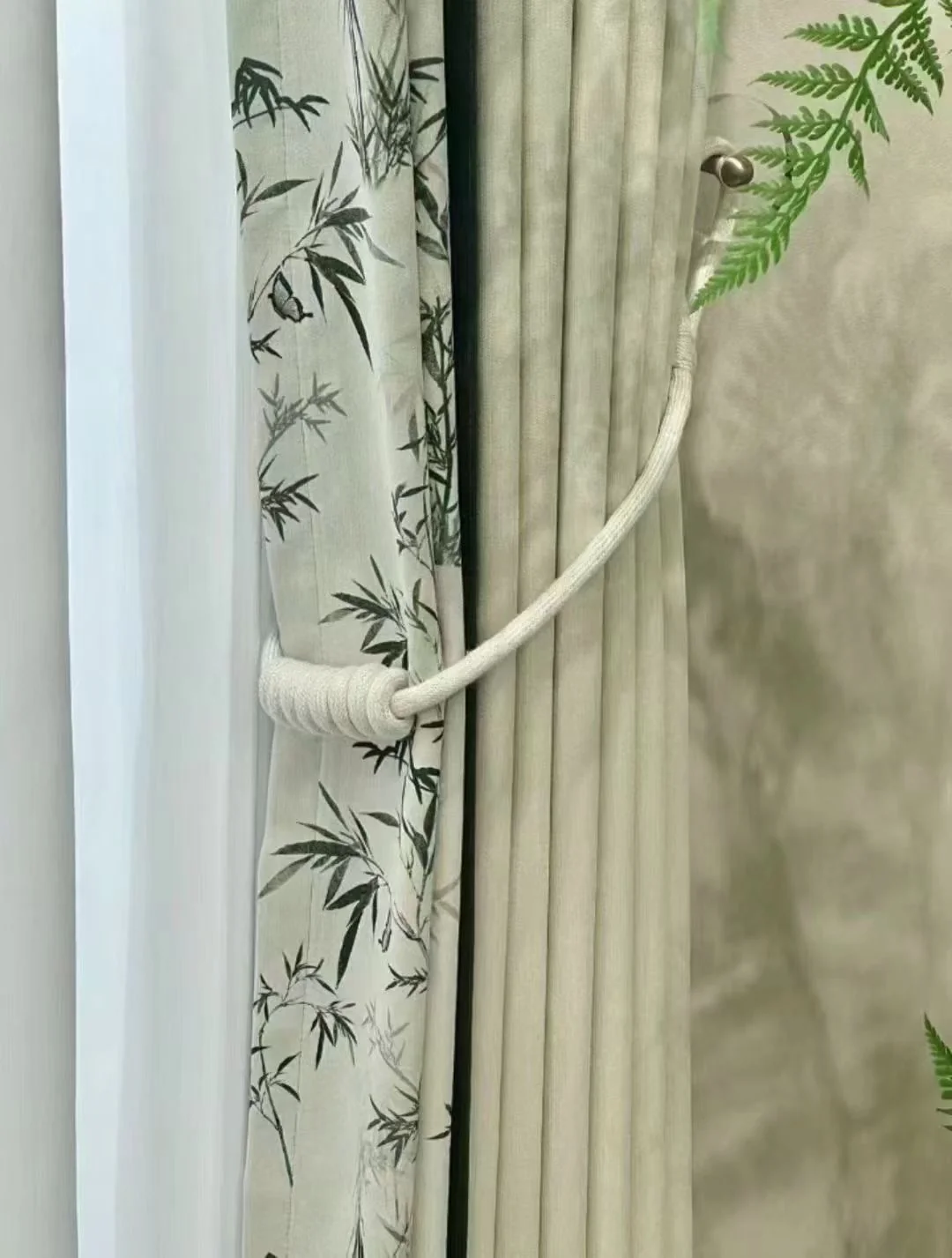 Bamboo leaf-patterned pale fabric beside a plain sage panel, held by curved light tieback.