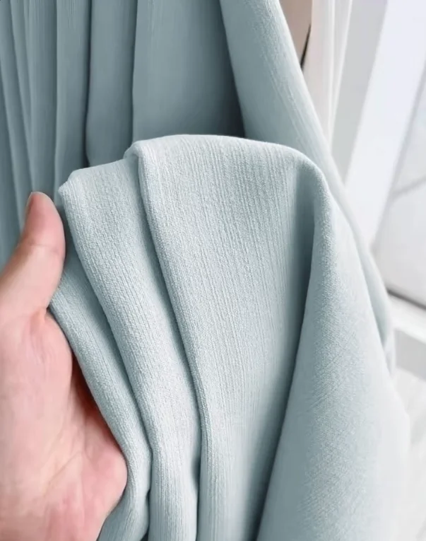 Hand holding powder-blue fabric against matching blue drapery, showing fine woven texture.