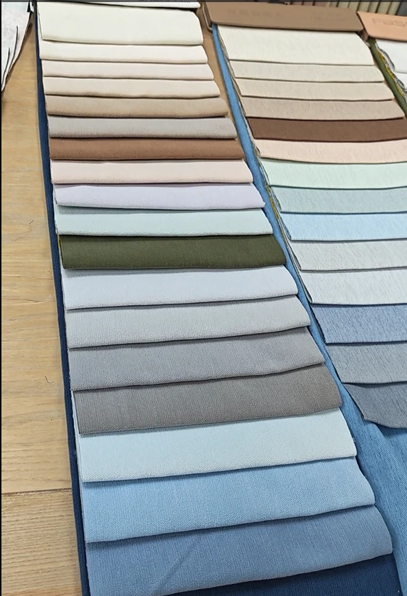 Night curtain fabric sample books displaying ivory, beige, taupe, grey, sage green and blue woven fabrics on a wooden table.