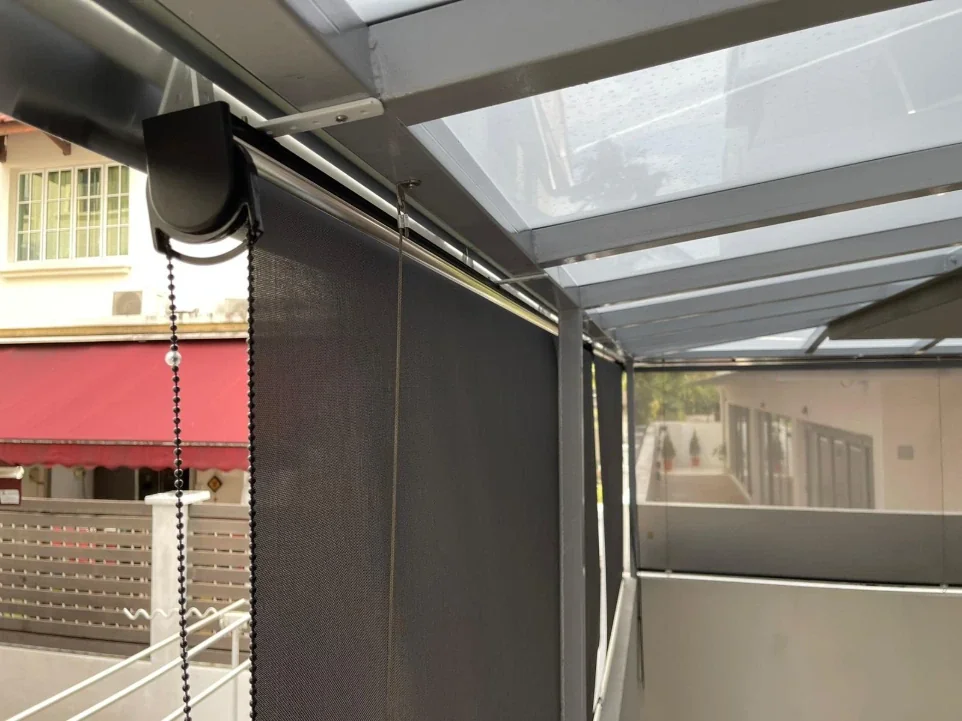 Outdoor roller screen under a glazed awning, with exposed roller tube, chain and guide/support hardware.