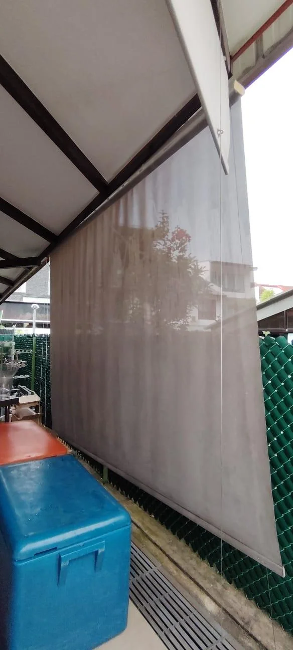 Long brown outdoor screen below a metal shelter beside storage tubs and fence; source placed under wire outdoor blinds.