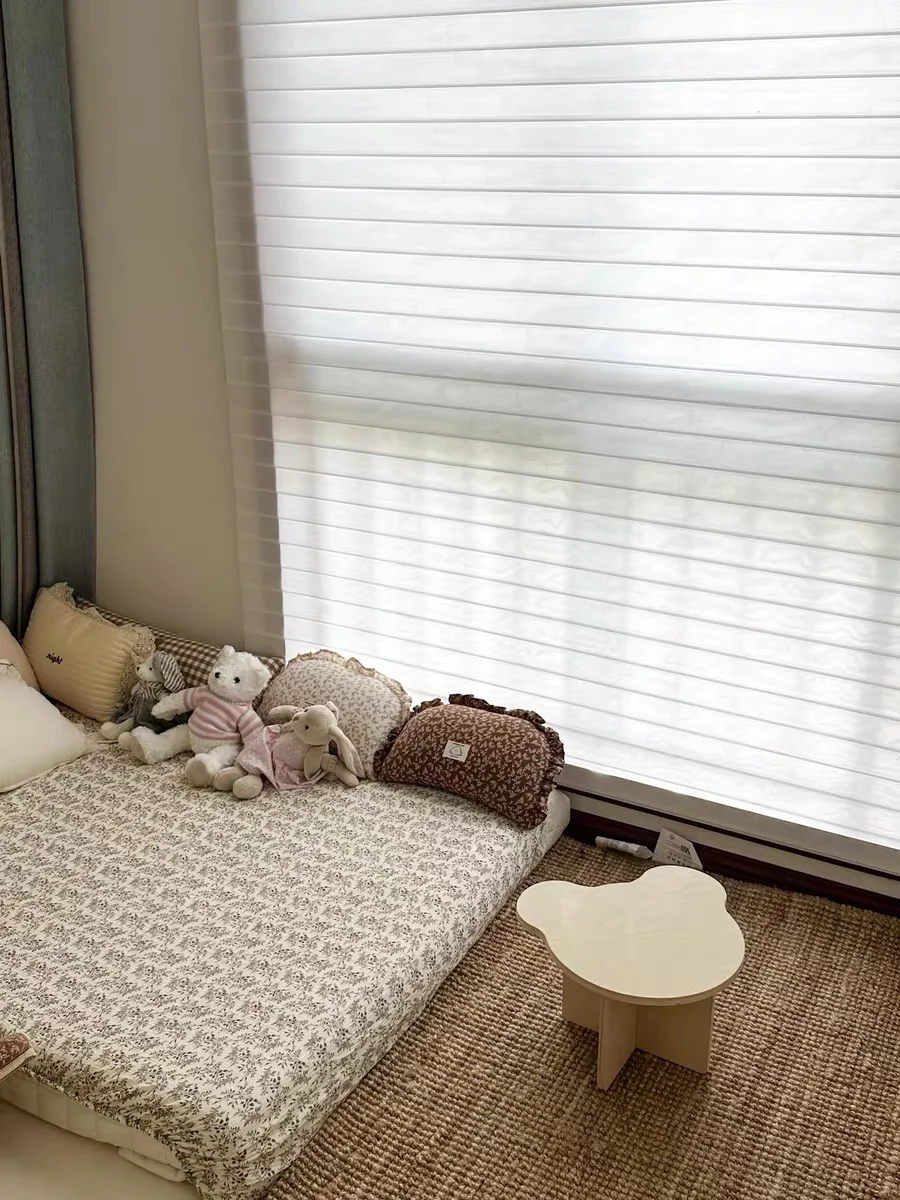 Bedroom window covered by a pale layered horizontal-vane sheer blind; bed and toys below.
