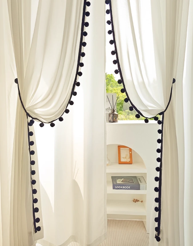 Close view of softly gathered ivory sheer curtains finished with contrasting black pom-pom trim along the curved and vertical edges.