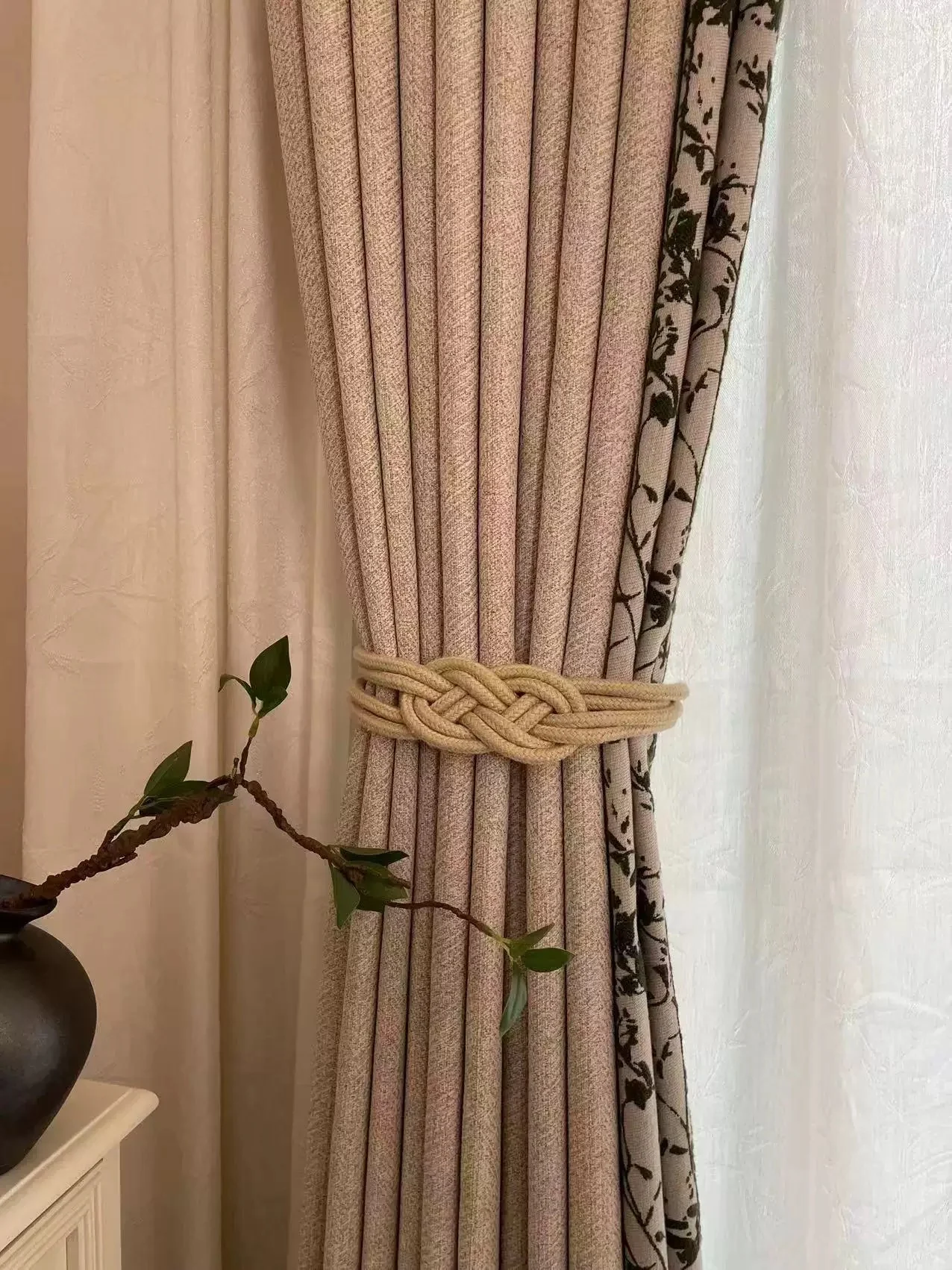 Warm oatmeal drape with contrasting botanical black vine/leaf strip at vertical leading edge, chunky cream rope knot tieback — supplied curtain design reference.