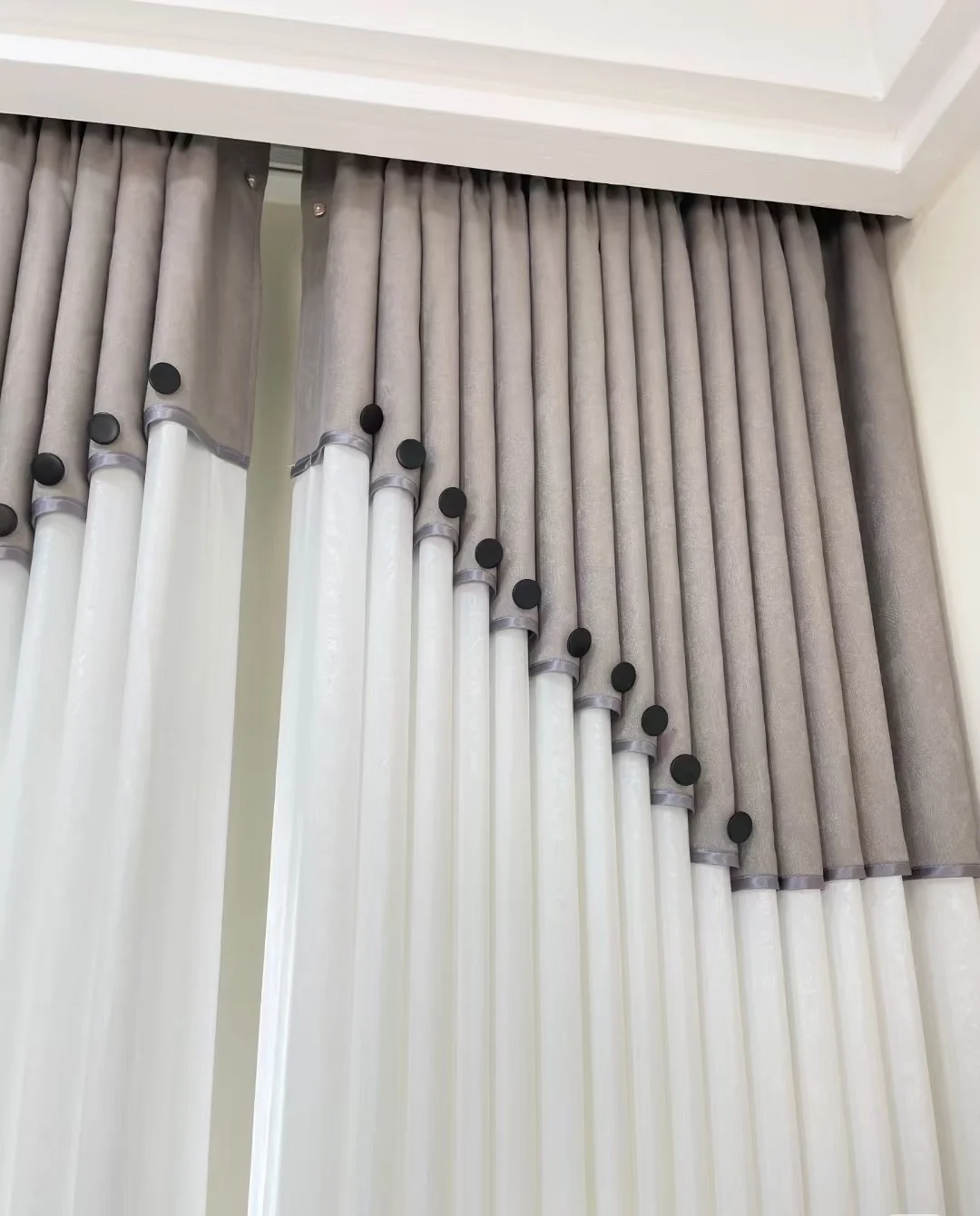 Grey-taupe upper curtain section with diagonal scalloped boundary formed across folds, small matte black disc accents and slim grey piping above flowing white sheer lower area — supplied curtain design reference.