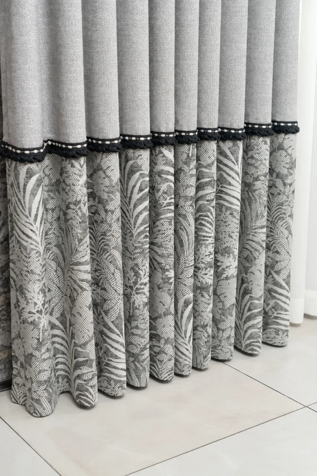 Silver-grey plain upper curtain, broad grey botanical leaf-pattern lower quarter, narrow black-and-ivory braid at horizontal join — supplied curtain design reference.