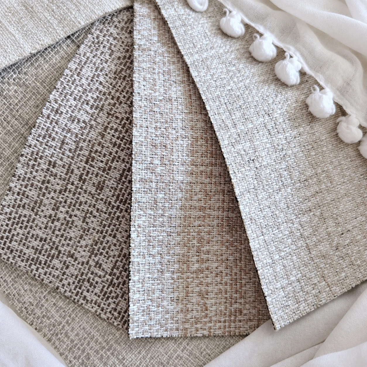 Cream, taupe and silver-grey crosswoven fabric samples with white pom-pom sheer trim.