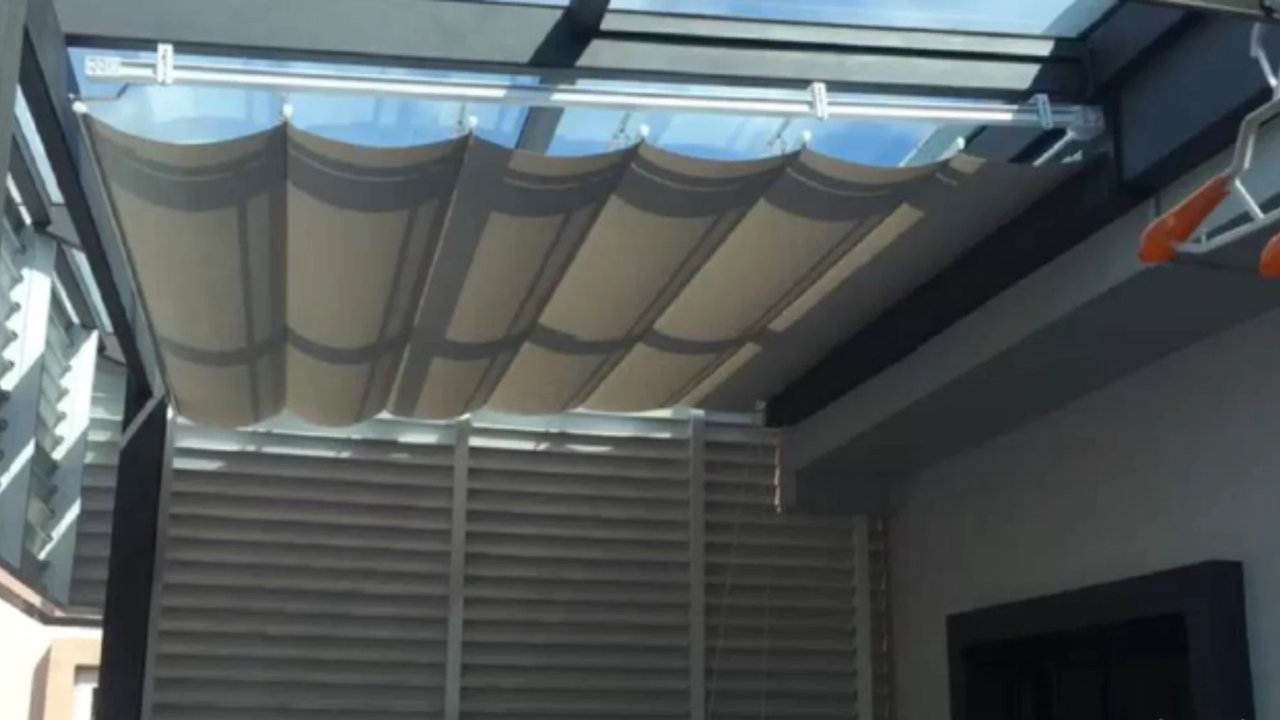 Folded pale fabric canopy spanning overhead tracks beneath a dark-framed glass roof, with uncovered glazing visible above.