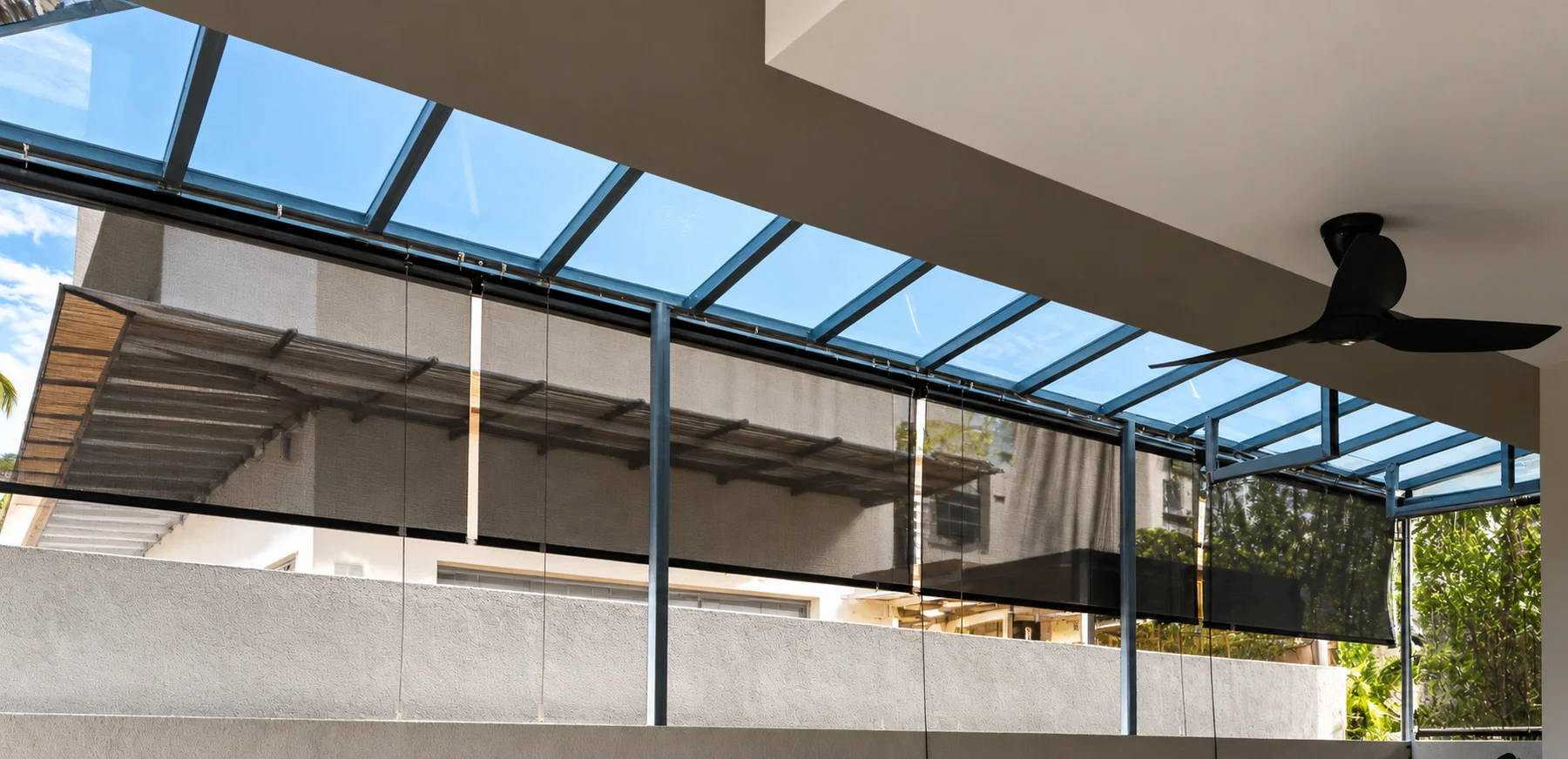 AI-restored source view of brown cable-guided outdoor roller screens beneath a blue-grey glazed patio roof, with boundary wall and ceiling fan.