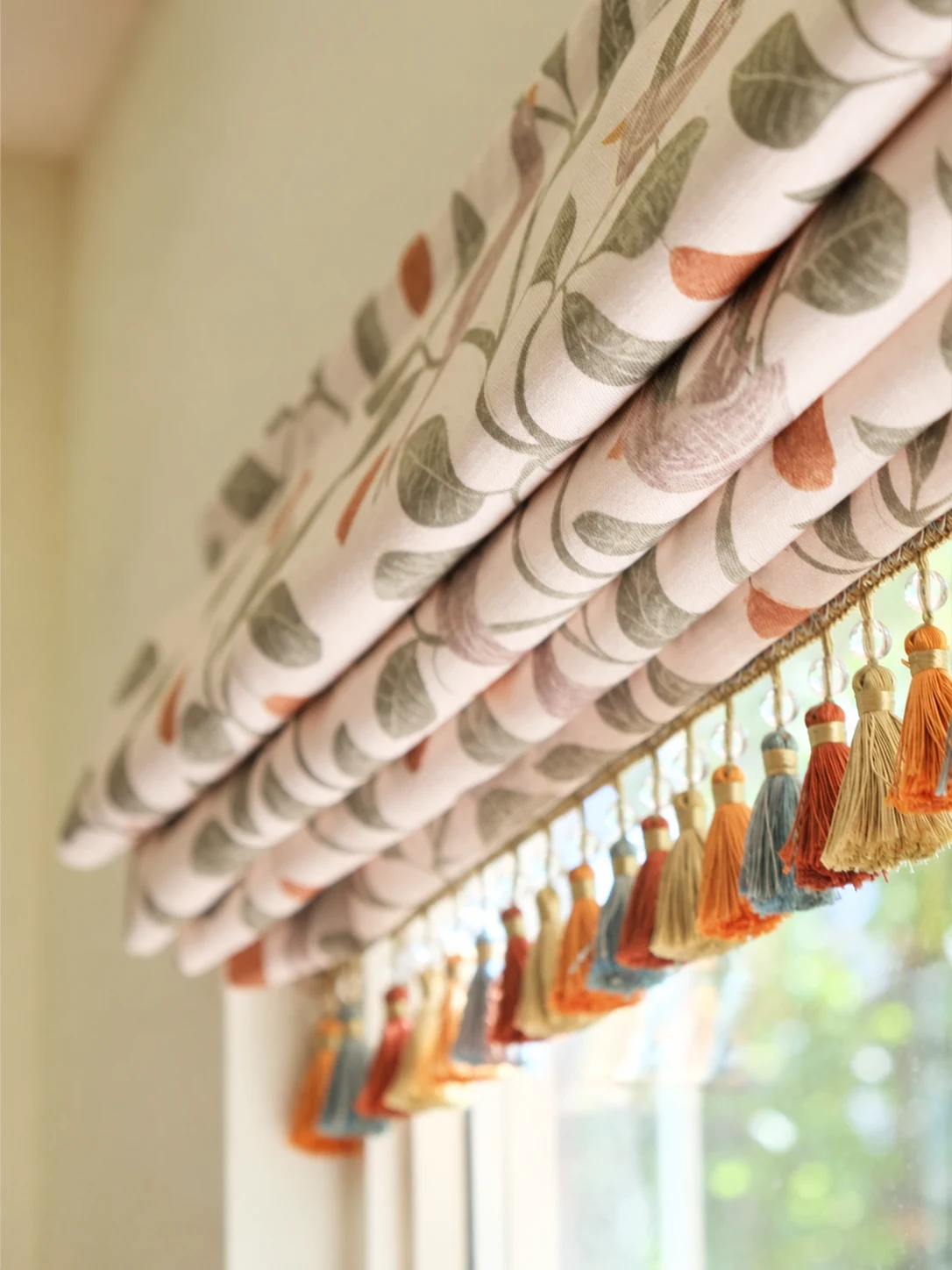 Fabric Roman shade with stacked horizontal folds, patterned botanical fabric and multicolour tassel trim.