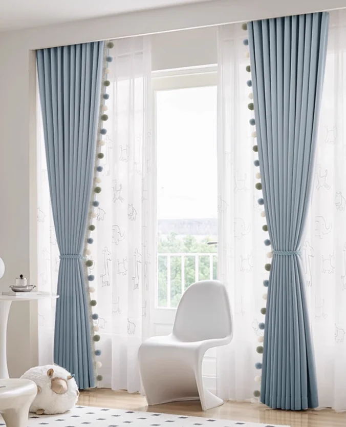 Blue night curtains with white elephant-patterned sheers and pom-pom trim in a child’s play and reading corner.